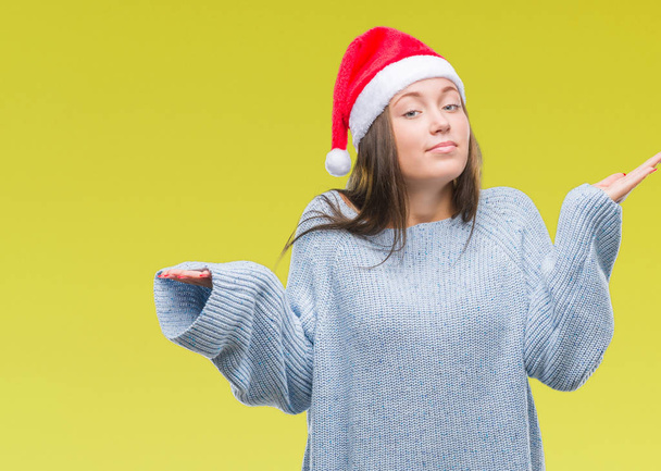 若い美しい白人女性の腕と手を上げて分離背景無知と混乱する式でクリスマス帽子をかぶっています。疑いのコンセプト. - 写真・画像