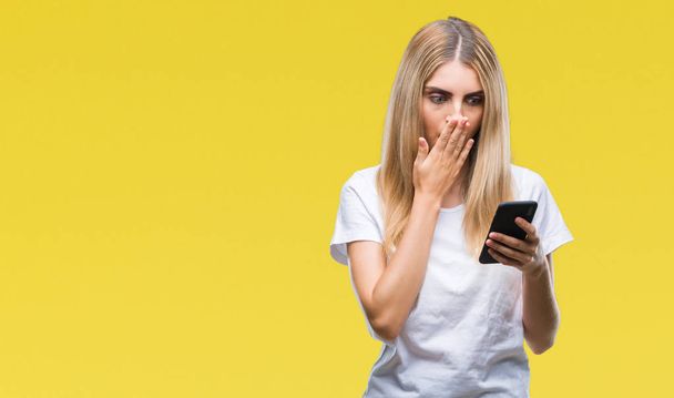 Young beautiful blonde woman using smartphone over isolated background cover mouth with hand shocked with shame for mistake, expression of fear, scared in silence, secret concept - Foto, Imagem