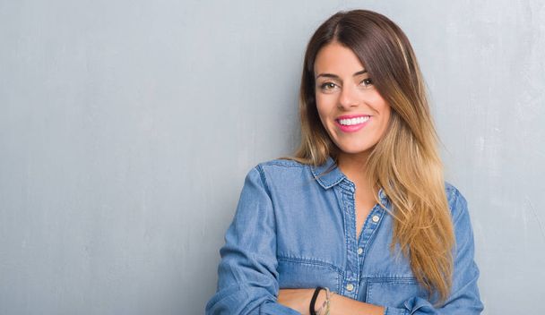 Young adult woman over grunge grey wall wearing denim outfit happy face smiling with crossed arms looking at the camera. Positive person. - Photo, Image