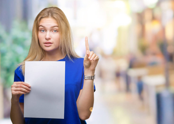 junge kaukasische Frau hält leeres Papierblatt über isoliertem Hintergrund und überrascht mit einer Idee oder Frage, die mit glücklichem Gesicht zeigt, Nummer eins - Foto, Bild