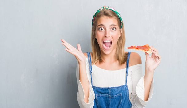Gyönyörű fiatal nő, mint a grunge szürke falon eszik pepperoni pizza szelet, nagyon boldog és izgatott, győztes kifejezés ünneplése győzelem nagy mosollyal sikoltozik, és kezet emelt - Fotó, kép