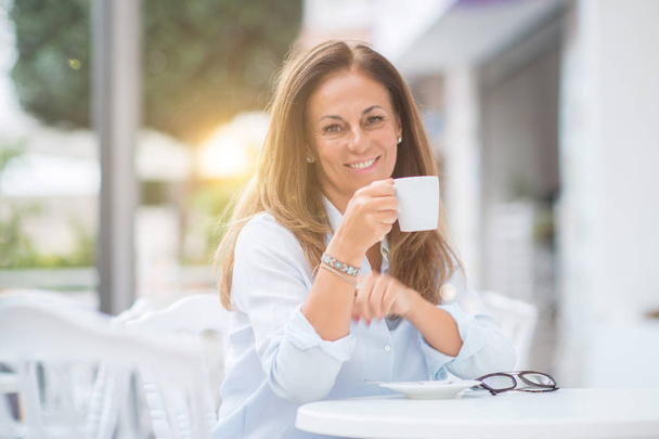 Hermosa mujer hispana de mediana edad en la cafetería con sonrisa en la cara en la ciudad. Beber una taza de café seguro y alegre en la terraza en un día soleado
. - Foto, imagen