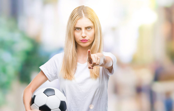 Молодая красивая блондинка держит футбольный мяч на изолированном фоне указывая пальцем на камеру и на вас, знак руки, позитивный и уверенный жест спереди
 - Фото, изображение