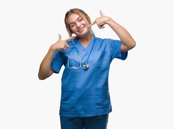 Fiatal kaukázusi doktor nő visel sebész egységes, több mint elszigetelt háttér mosolyogva magabiztos bemutatás és mutat ujjak fogak és a száj. Egészségügyi koncepció. - Fotó, kép