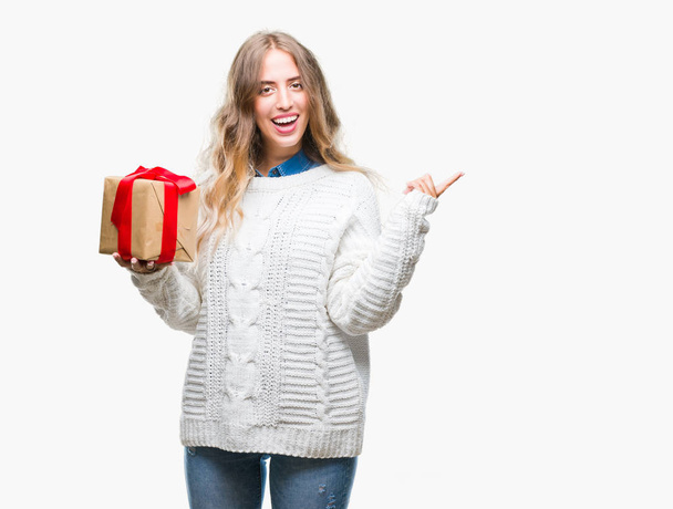 Beautiful young blonde woman holding gift over isolated background very happy pointing with hand and finger to the side - Fotó, kép