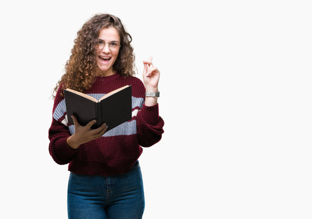 Mladá brunetka s knihou nošení brýlí izolované pozadí překvapilo s nápad nebo otázku ukazující prst s šťastné tváře, číslo jedna - Fotografie, Obrázek