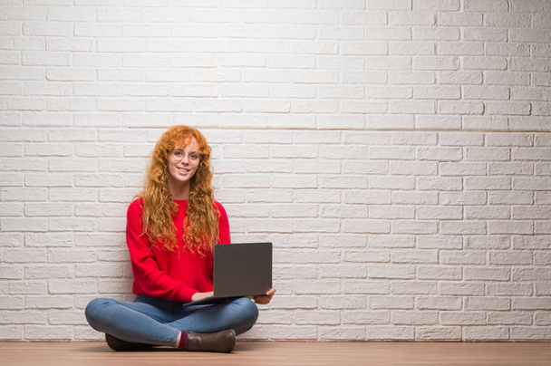 Young redhead woman sitting over brick wall using computer laptop with a happy face standing and smiling with a confident smile showing teeth - Photo, Image
