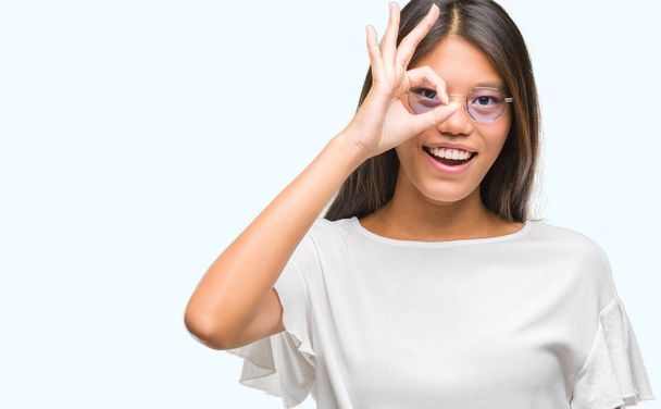 Νέοι Ασιατικές γυναίκα φοράει γυαλιά, πέρα από το απομονωμένο υπόβαθρο κάνει εντάξει χειρονομία με το χέρι, χαμογελώντας, το μάτι ψάχνει μέσα από τα δάχτυλα με χαρούμενο πρόσωπο. - Φωτογραφία, εικόνα