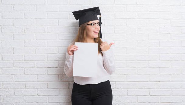 Молода доросла жінка над білою цегляною стіною в шапці випускника, що тримає ступінь, вказуючи і показуючи з великим пальцем в сторону з щасливим обличчям посміхаючись
 - Фото, зображення