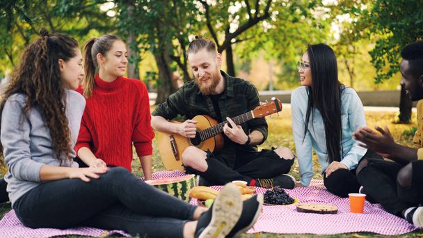 Joyful hipster is singing and playing the guitar sitting on blanket in park with friends and having fun, people are clapping hands. Food, music and fun concept. - Photo, Image
