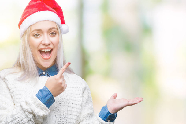 分離背景驚くし、手とを提示、指で指している間カメラに笑顔でクリスマス帽子身に着けている若いブロンドの女性. - 写真・画像