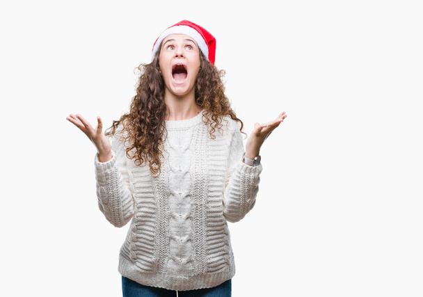 Mladá bruneta dívka, která nosí vánoční čepice nad izolované pozadí blázen a mad křičet a řvát s agresivní výraz a zvedl ruce. Frustrace koncept. - Fotografie, Obrázek
