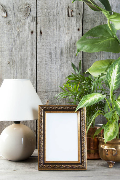 Üres sárgaréz függőleges képkeret, zöld növények és egy asztali lámpa a régi fa szürke textúrázott háttérre. Otthoni dekoráció és másol adható szöveg.   - Fotó, kép