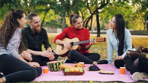 Charming young woman is playing the guitar sitting on blanket with friends on picnic, girls and guys are clapping hands and listening to music. Fun and nature concept. - Fotografie, Obrázek
