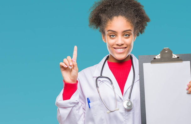 Jovem afro-americana médico mulher segurando prancheta sobre fundo isolado surpreendido com uma ideia ou pergunta apontando dedo com cara feliz, número um
 - Foto, Imagem