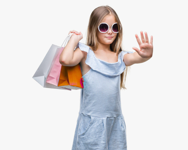 Young beautiful girl holding shopping bags on sales over isolated background with open hand doing stop sign with serious and confident expression, defense gesture - Photo, Image