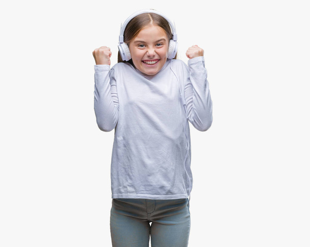 Όμορφη κοπέλα φορώντας ακουστικά να ακούτε μουσική μέσω απομονωμένες φόντο γιορτάζει έκπληκτος και κατάπληκτος για την επιτυχία με τα χέρια υψωμένα και ανοίξουν τα μάτια. Νικητής έννοια. - Φωτογραφία, εικόνα