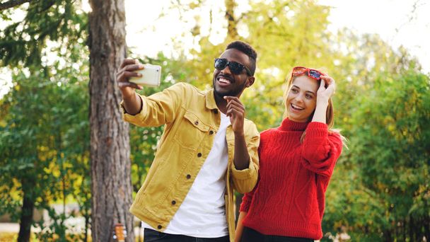 African American guy is taking selfie with female friend holding smartphone and posing with sunglasses standing in park together. Friendship and modern technology concept. - 写真・画像