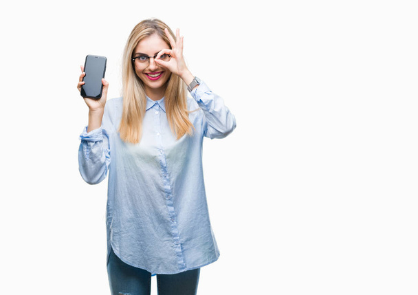 Young beautiful blonde business woman showing screen of smartphone over isolated background with happy face smiling doing ok sign with hand on eye looking through fingers - Foto, Imagem