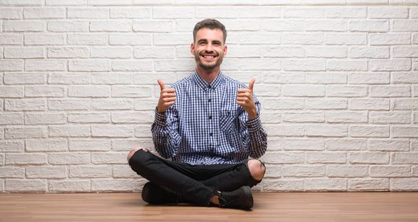 Hombre adulto joven sentado sobre la pared de ladrillo blanco signo de éxito haciendo gesto positivo con la mano, pulgares hacia arriba sonriendo y feliz. Mirando a la cámara con expresión alegre, gesto ganador
. - Foto, imagen