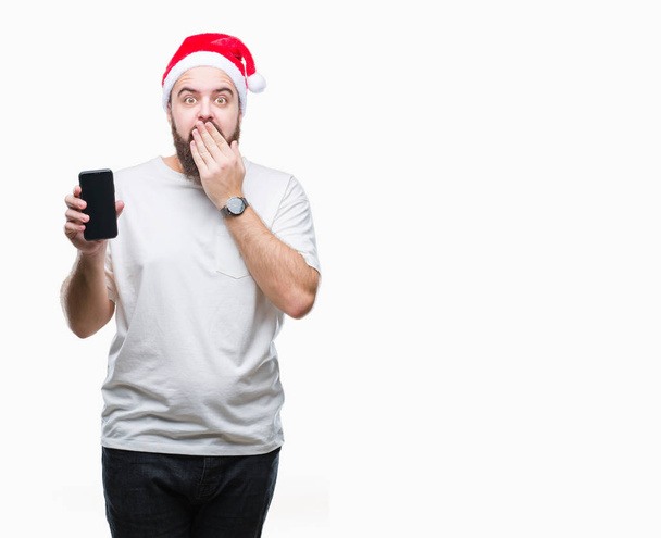 Młody człowiek kaukaski noszenia Boże Narodzenie ekranu smartfonu wyświetlone na kapelusz na białym tle okładka usta ręką w szoku ze wstydem za błąd, wyrażenie strachu, przestraszony w milczeniu, tajne koncepcja - Zdjęcie, obraz