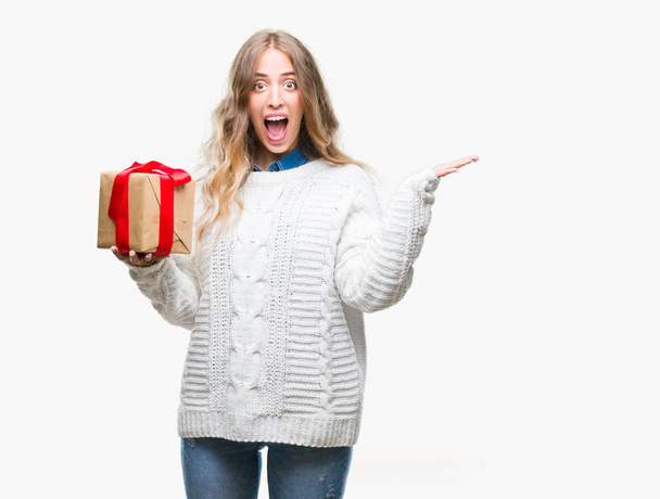 Beautiful young blonde woman holding gift over isolated background very happy and excited, winner expression celebrating victory screaming with big smile and raised hands - Фото, зображення