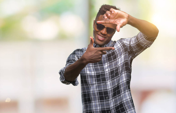 Młody człowiek african american, noszenie okularów przeciwsłonecznych na białym tle uśmiechający się podejmowania ramki z rąk i palców z szczęśliwy twarz. Koncepcja kreatywność i fotografii. - Zdjęcie, obraz