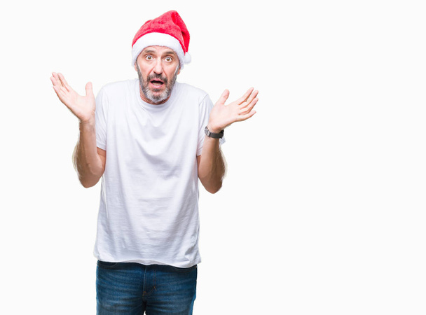 中年白髪年配の男性にクリスマスの帽子をかぶっては、腕と手を上げて背景無知と混乱式を分離しました。疑いのコンセプト. - 写真・画像