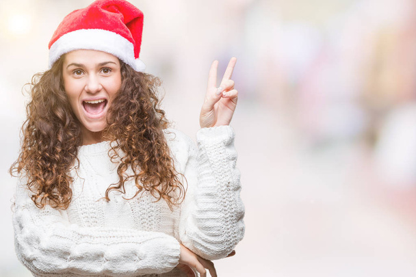 若いブルネットの少女は勝利のサインを行うカメラでウィンクしている幸せそうな顔を浮かべて隔離された背景にクリスマス帽子をかぶっています。2 位. - 写真・画像