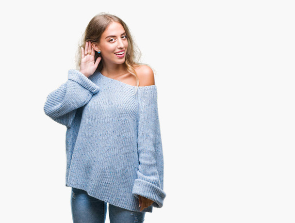 Beautiful young blonde woman wearing winter sweater over isolated background smiling with hand over ear listening an hearing to rumor or gossip. Deafness concept. - Photo, image