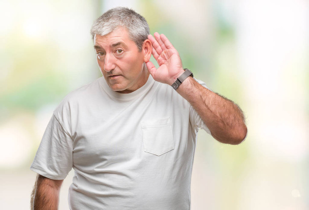 Pěkný starší muž izolované pozadí s úsměvem s rukou přes ucho naslouchá slyšení zvěsti nebo drby. Hluchota koncept. - Fotografie, Obrázek