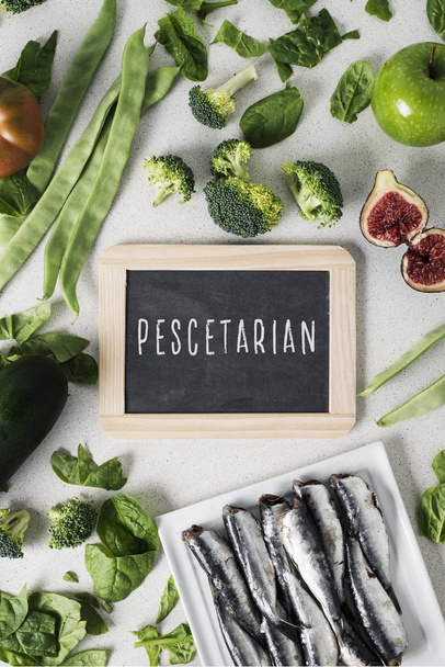 魚やいくつかのさまざまな果物や生の野菜の山を食べるベジタリアンの人のためのテキスト pescetarian で看板と白い石のカウンター上の皿にイワシの山 - 写真・画像