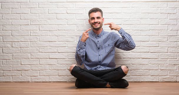 Homem adulto jovem sentado sobre a parede de tijolo branco sorrindo mostrando confiante e apontando com os dedos dentes e boca. Conceito de saúde
. - Foto, Imagem
