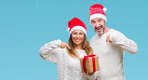 Hispanisches Paar mittleren Alters trägt Weihnachtsmütze und hält Geschenk über isoliertem Hintergrund mit Überraschungsgesicht, das mit dem Finger auf sich selbst zeigt - Foto, Bild