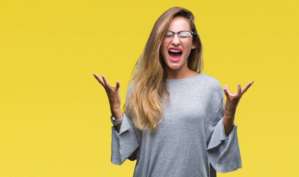 Young beautiful blonde woman wearing glasses over isolated background celebrating mad and crazy for success with arms raised and closed eyes screaming excited. Winner concept - Fotografie, Obrázek