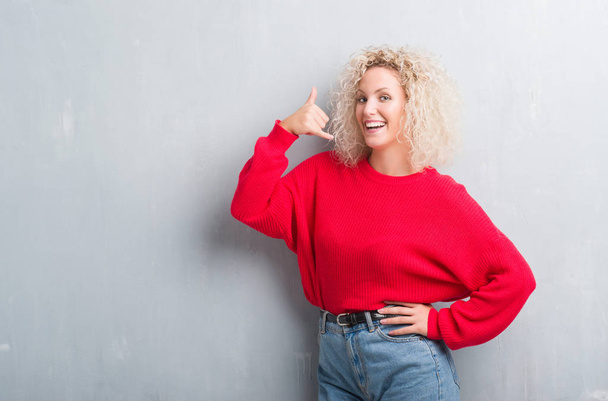 Young blonde woman with curly hair over grunge grey background smiling doing phone gesture with hand and fingers like talking on the telephone. Communicating concepts. - Photo, Image