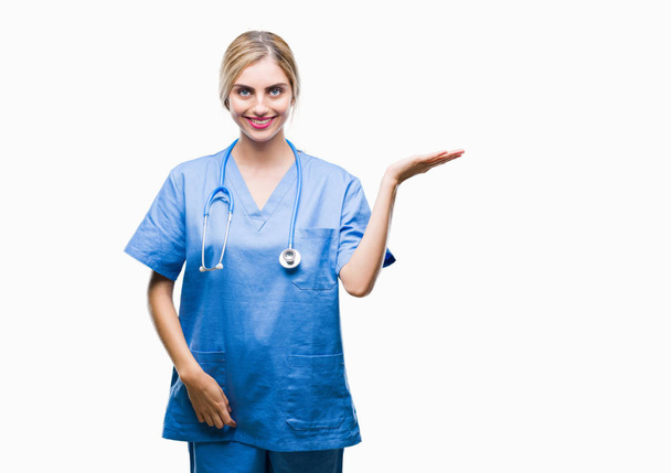 Молода красива блондинка лікар хірург медсестра на ізольованому фоні усміхнений веселий подарунок і вказує рукою на камеру
. - Фото, зображення
