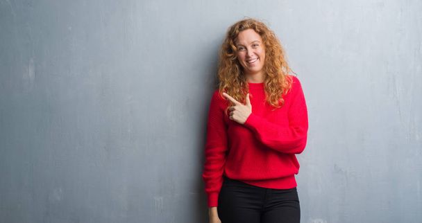 Mujer pelirroja joven sobre la pared gris grunge vistiendo suéter rojo alegre con una sonrisa de la cara apuntando con la mano y el dedo hacia el lado con expresión feliz y natural en la cara mirando a la cámara
. - Foto, Imagen