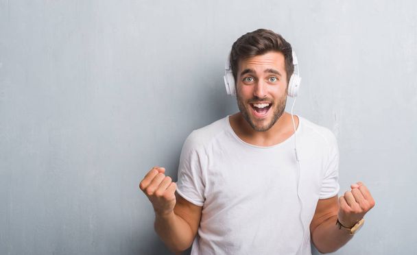 Handsome young man over grey grunge wall wearing headphones listening to music screaming proud and celebrating victory and success very excited, cheering emotion - Photo, Image