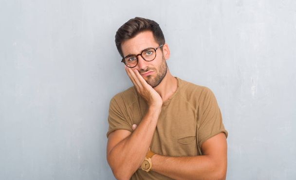 Knappe jonge man over grijs grunge muur dragen bril denken op zoek moe en verveeld met depressie problemen met gekruiste armen. - Foto, afbeelding