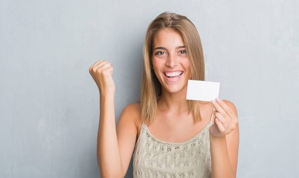 Beautiful young woman over grunge grey wall holding blank visit card screaming proud and celebrating victory and success very excited, cheering emotion - Foto, immagini