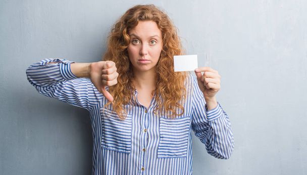 Молодая рыжая женщина над серой гранж-стеной держит чистую визитную карточку с сердитым лицом, негативный знак показывает неприязнь с опущенными пальцами, отвержение концепции
 - Фото, изображение