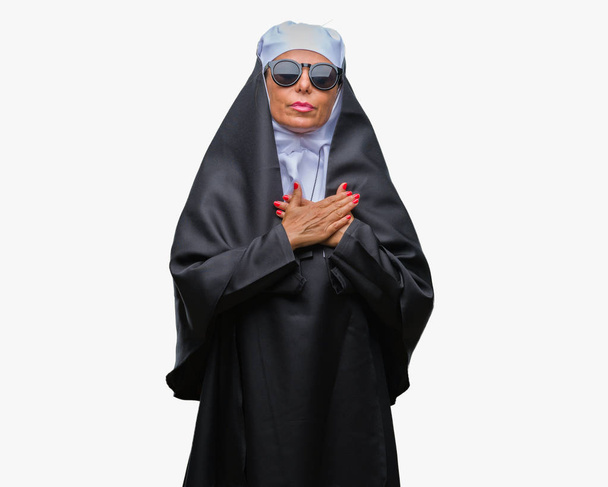 Femme religieuse catholique âgée du Moyen Âge portant des lunettes de soleil sur fond isolé souriant avec les mains sur la poitrine avec les yeux fermés et geste reconnaissant sur le visage. Concept de santé
. - Photo, image