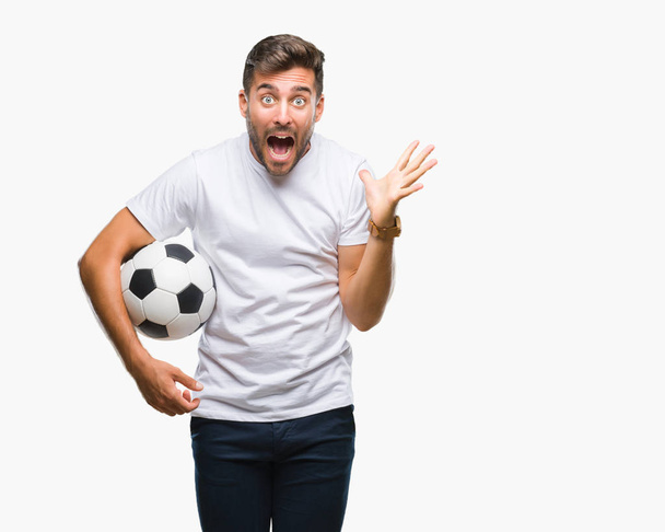 Joven hombre guapo sosteniendo pelota de fútbol sobre un fondo aislado muy feliz y emocionado, expresión ganadora celebrando la victoria gritando con una gran sonrisa y levantando las manos
 - Foto, imagen