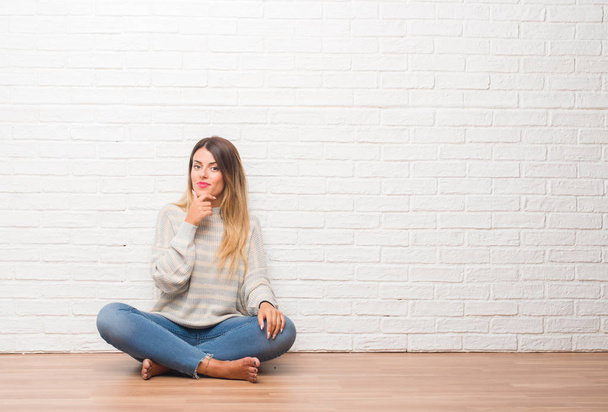Νεαρή ενήλικη γυναίκα που κάθεται στο πάτωμα πάνω από το λευκό τοίχο στο σπίτι αναζητούν αυτοπεποίθηση στη φωτογραφική μηχανή με χαμόγελο με σταυρωμένα χέρια και χέρι σηκωμένο στο πηγούνι. Σκέφτεστε θετικά. - Φωτογραφία, εικόνα