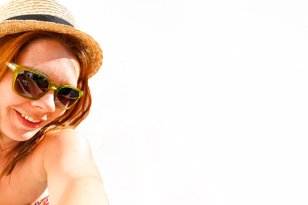 Young smiling red-haired woman in a straw hat and sunglasses photographing a selfie, isolated on white background. - Photo, Image