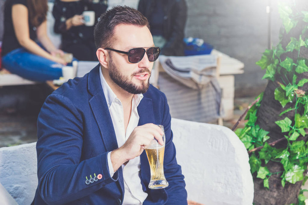 Portrait of fashionable man sitting in cafe pub outdoors and holding glass of beer. - Photo, image