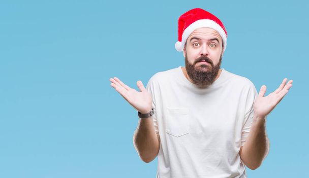 若い白人ヒップ男は腕と手を上げて分離背景無知と混乱する式でクリスマス帽子をかぶっています。疑いのコンセプト. - 写真・画像