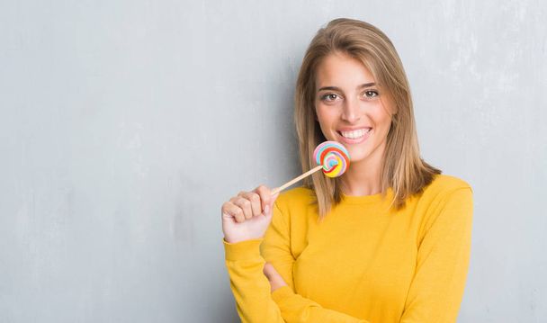 Bella giovane donna sopra grunge muro grigio mangiare caramelle lecca-lecca con una faccia felice in piedi e sorridente con un sorriso fiducioso che mostra i denti
 - Foto, immagini