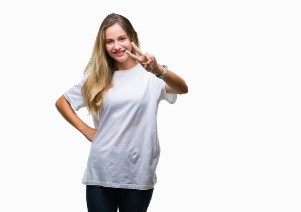 Fiatal gyönyörű szőke nő visel alkalmi fehér póló, mosolyogva, keres, hogy a kamera mutatja a győzelem jele ennek ujjak elszigetelt háttérhez képest. Két szám. - Fotó, kép